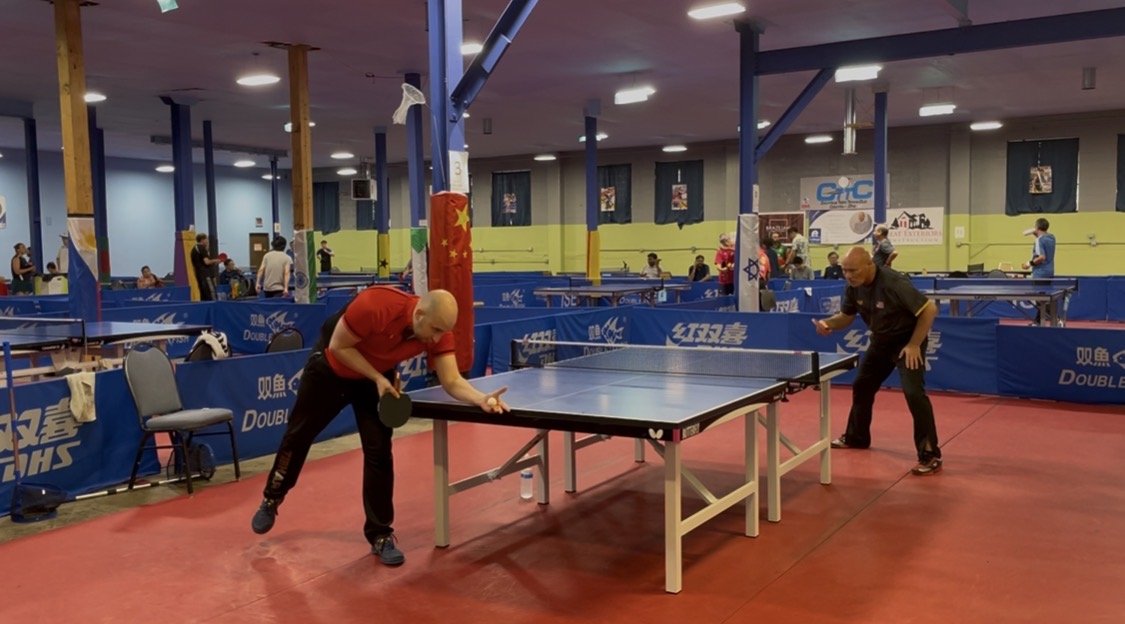 Two men playing table tennis in an indoor sports facility with multiple tables and people in the background.