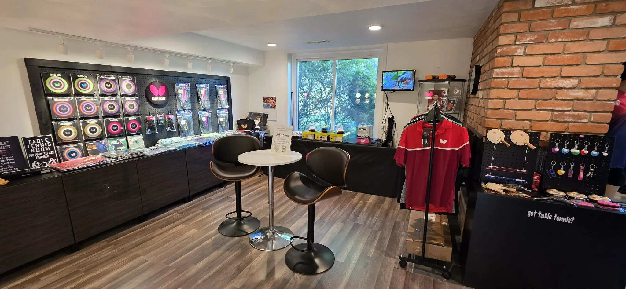 Indoor store with table tennis merchandise including paddles, balls, and accessories displayed on black shelves and pegboards. The room has a large window, a small round table with two chairs, and promotional signs, including one for table tennis.