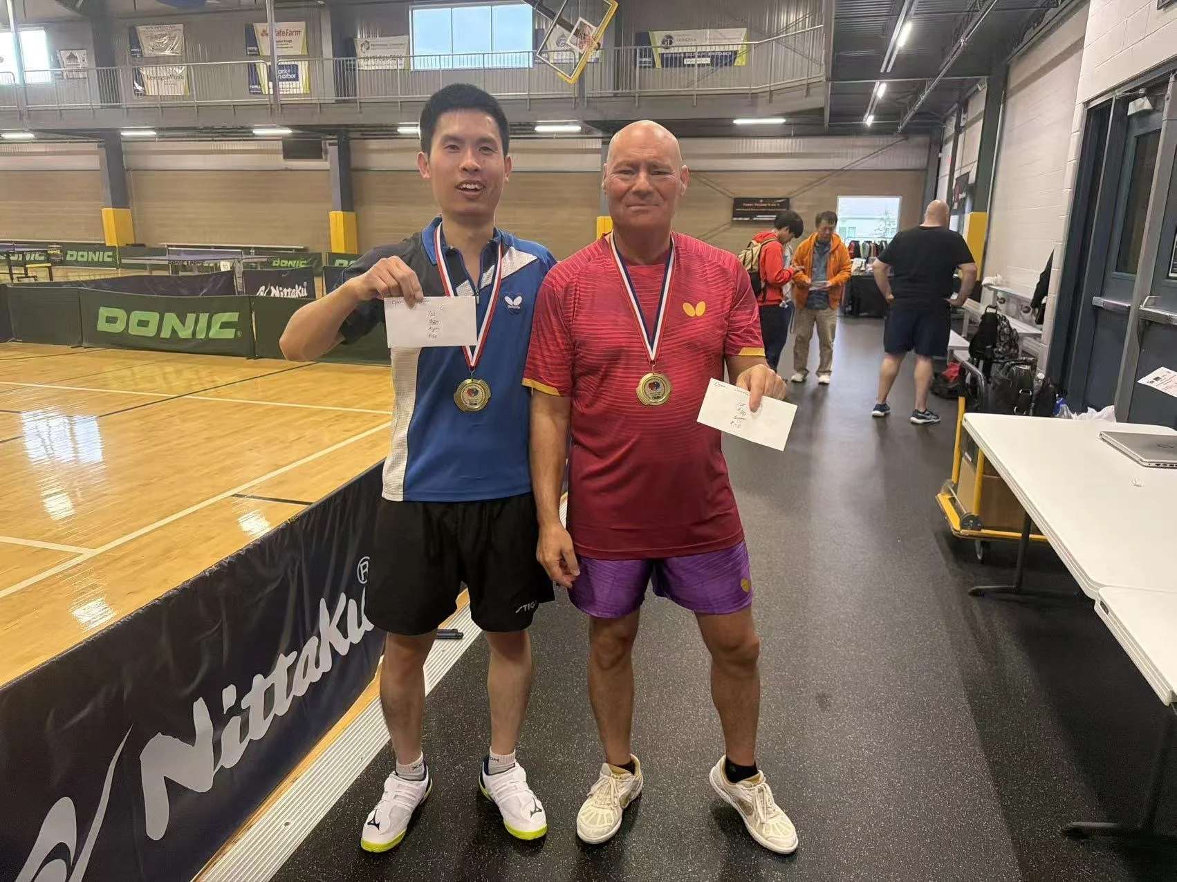 Two male table tennis players standing in an indoor sports facility, each wearing a medal around their neck and holding a piece of paper, smiling at the camera. The player on the left is in a blue and white shirt with black shorts and white sports sh