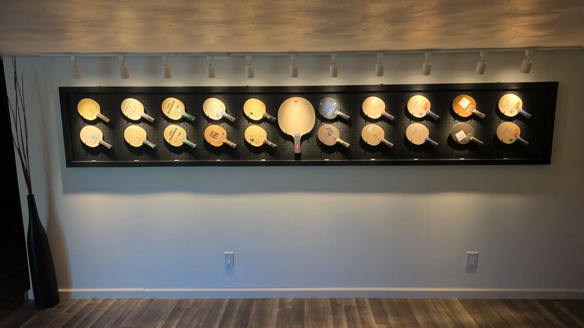 A display of multiple ping pong paddles mounted on a black pegboard wall, illuminated by ceiling spotlights in a retail store.