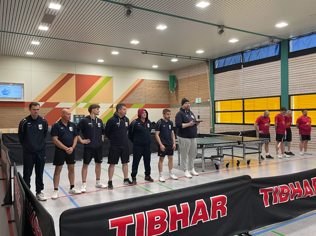 A group of people standing in a line on an indoor sports court, with some in sports attire and others in casual clothing, near a ping pong table.