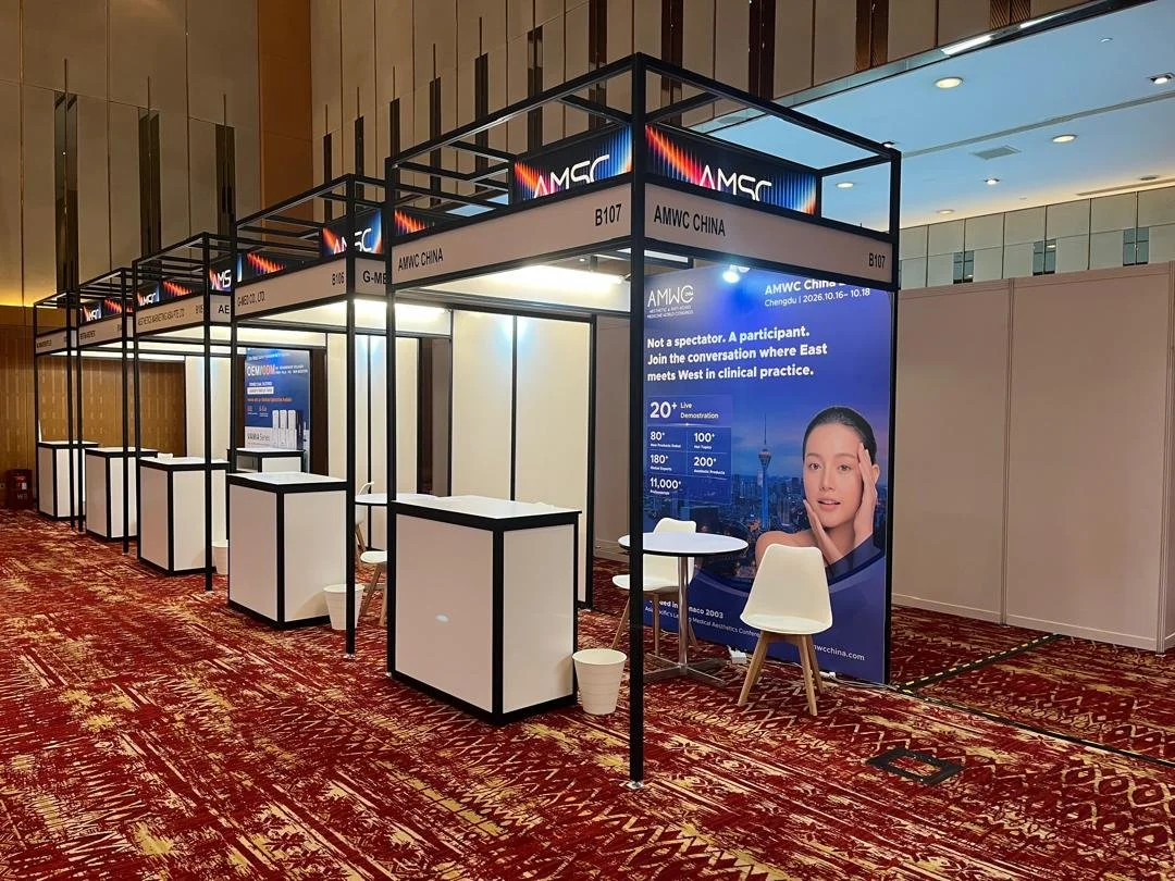 A large exhibition booth at a conference with the sign 'AMWC China' and a digital display promoting the event. The booth has multiple sections with white counters and chairs, set on a patterned red and yellow carpet.