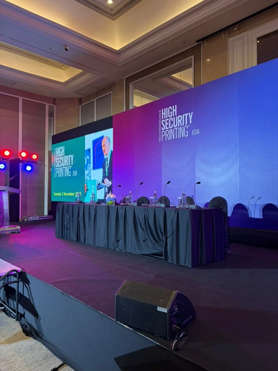 Conference stage with a large digital screen displaying the event title 'High Security Printing Asia' and a speaker presentation. The stage is set with a table, chairs, microphones, and water bottles, with colorful lighting in the background.