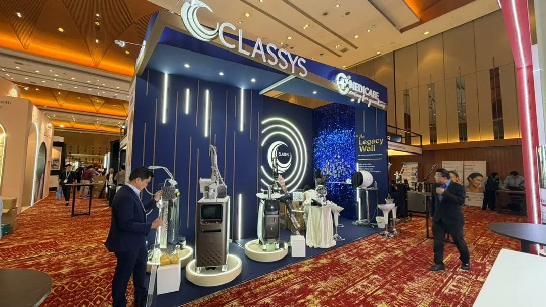 Trade show booth displaying medical equipment with the branding 'Classys' and 'Medicare'. The booth features advanced medical devices and a large illuminated sign, set in a convention center with red patterned carpet and gold ceiling accents.