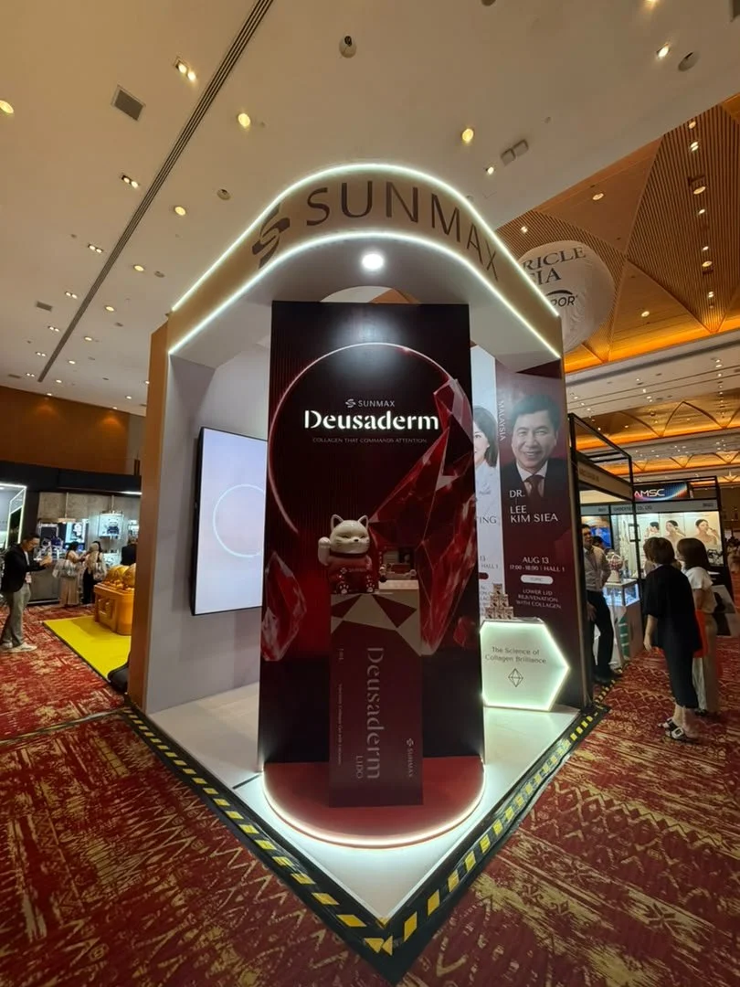 Exhibit at a trade show promoting Deusaderm, a collagen product by Sunmax, with a small white dog figurine on display, surrounded by banners and informational displays.