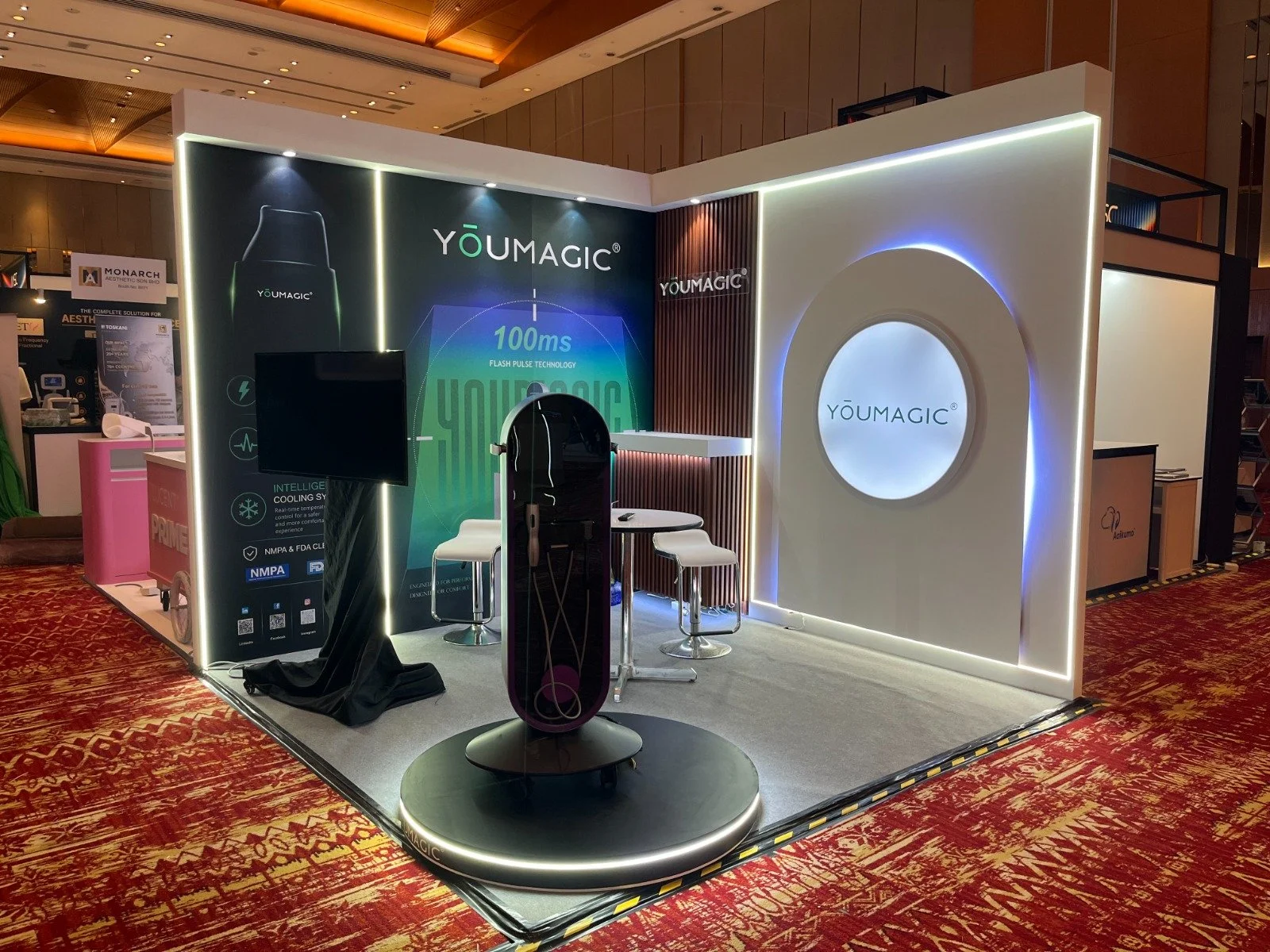 Exhibition booth for YouMagic showcasing advanced cooling technology with a circular display, seating, a monitor, and promotional banners in an event hall.