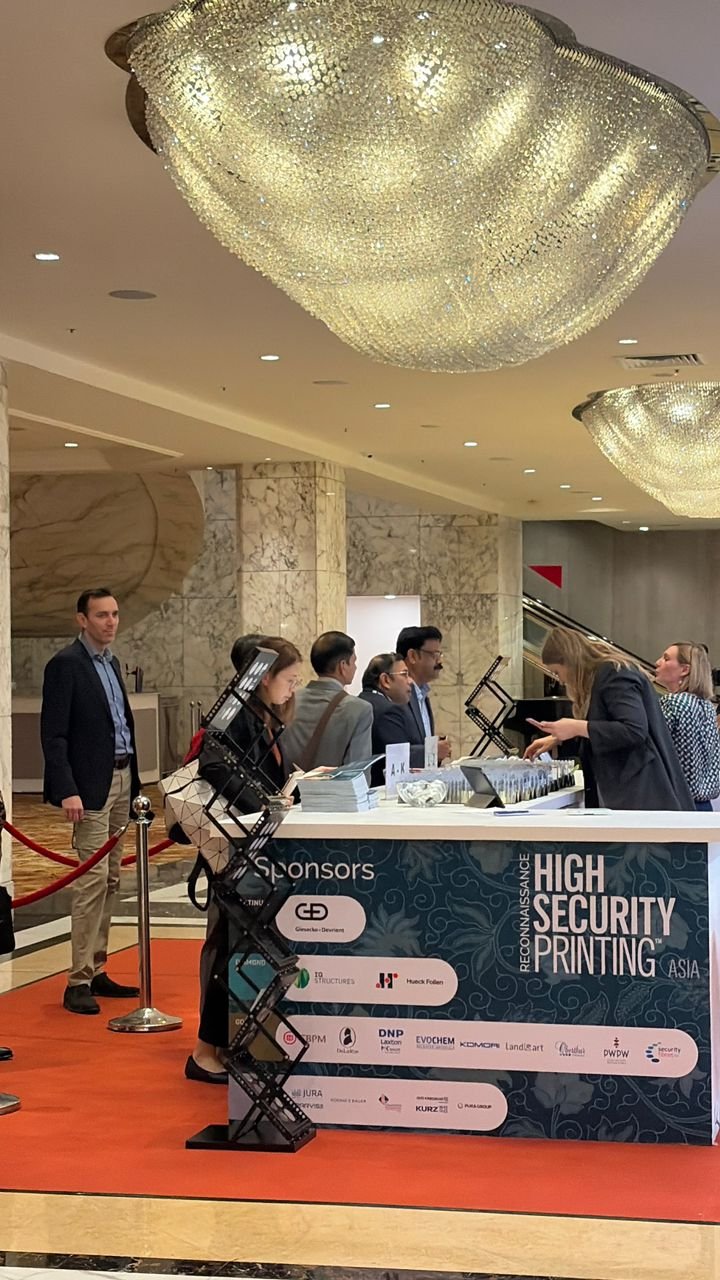 People standing and sitting at a high security printing event registration desk in a hotel lobby with large chandelier ceiling lights.