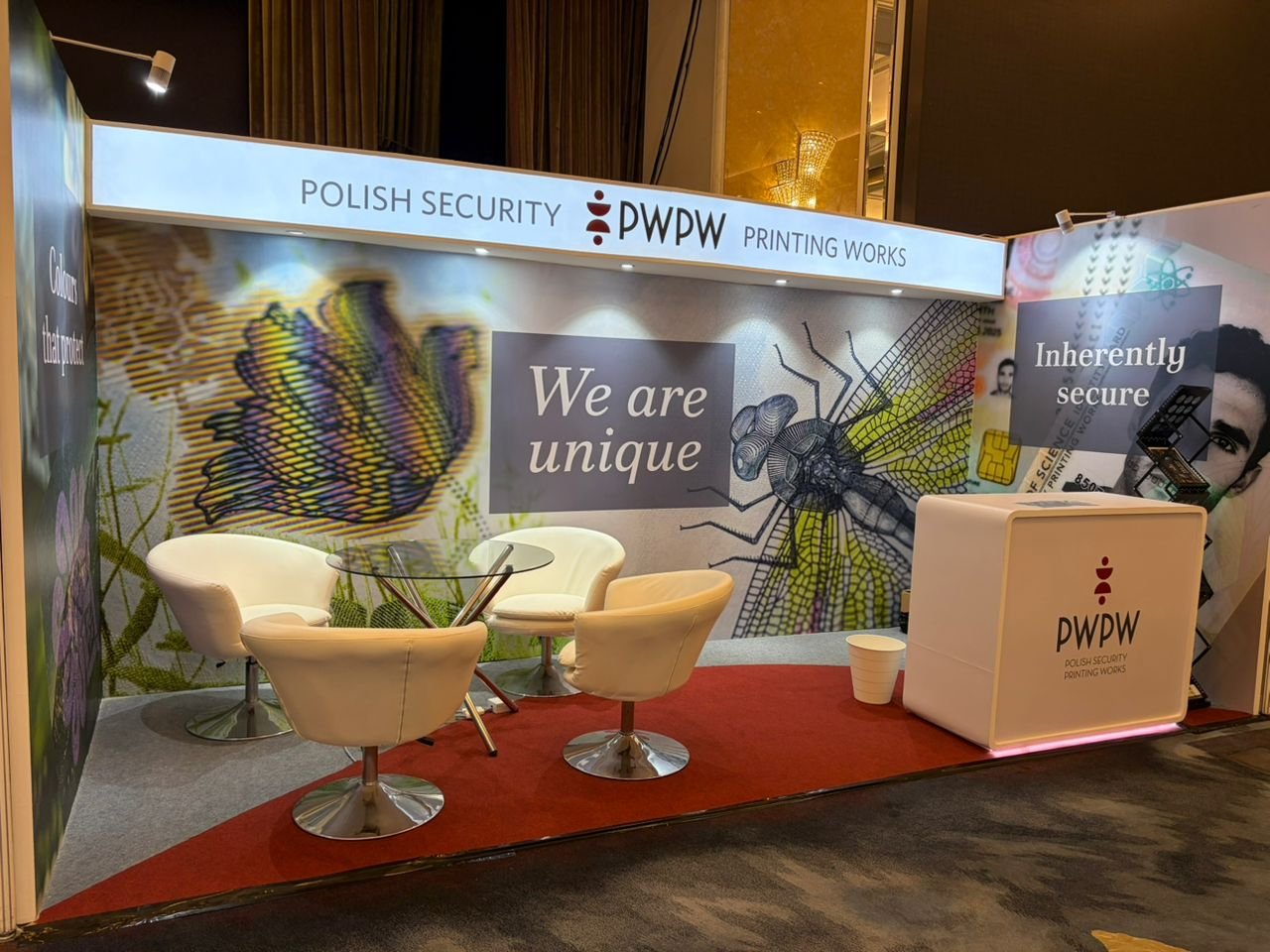 Exhibit booth for Polish Security Printing Works (PWPW), with a backdrop showing a colorful butterfly, a detailed insect illustration, and a person’s face on a poster. There are four white armchairs around a glass table on a red carpet, with a white counter displaying the PWPW logo and slogan.