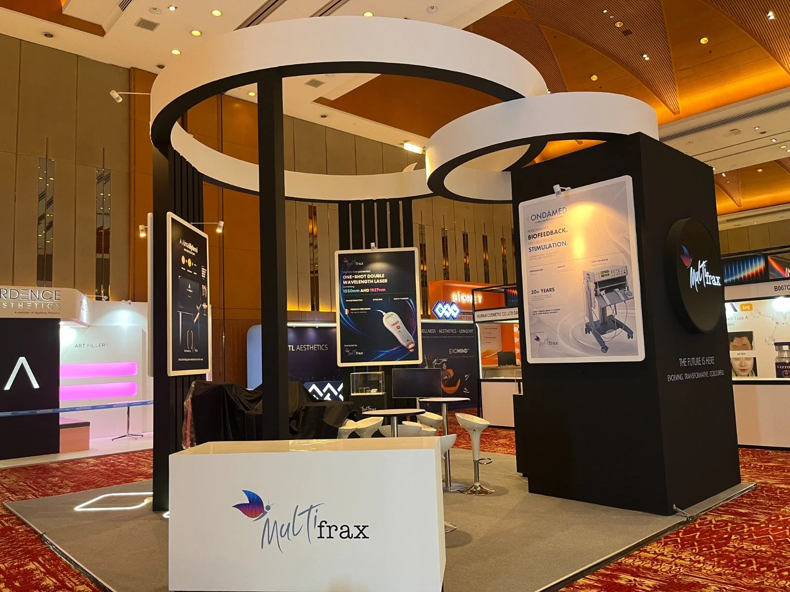 Trade show display with promotional posters and booths for aesthetic and medical products, featuring a large black and white circular structure and a black booth with the 'MultiTfrax' logo.