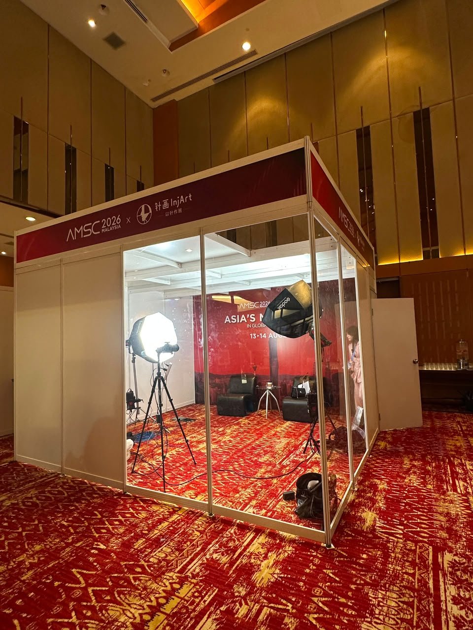 Photo of a conference booth with cameras and lighting equipment inside, on a red patterned carpet, at an event in Malaysia.