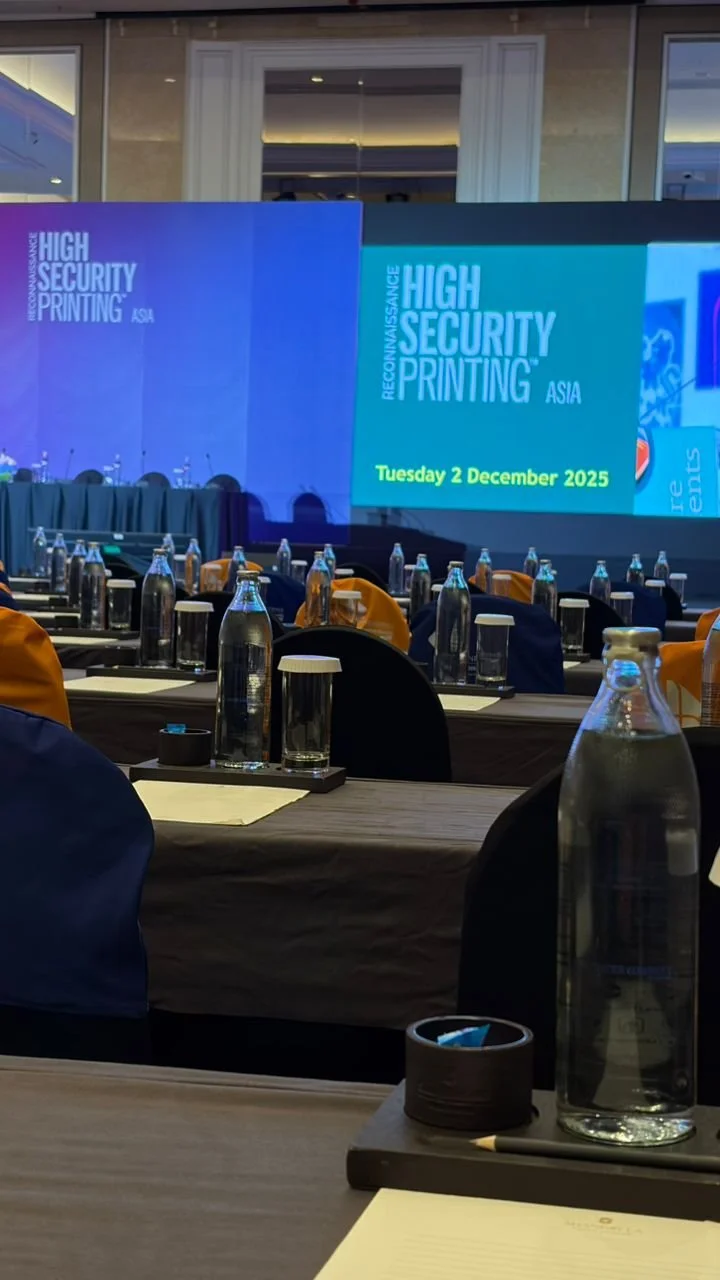 Conference room set up with rows of tables, water bottles, glasses, and notepads, facing a large screen displaying the event titled 'High Security Printing Asia' on December 2, 2025.