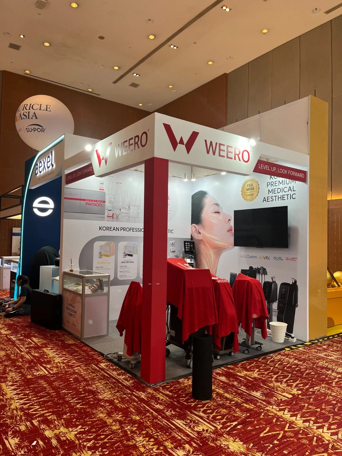 Trade show booth for Weero featuring skincare and medical aesthetic products, with a large poster of a woman's face and multiple product displays, set on a red and yellow patterned carpet.