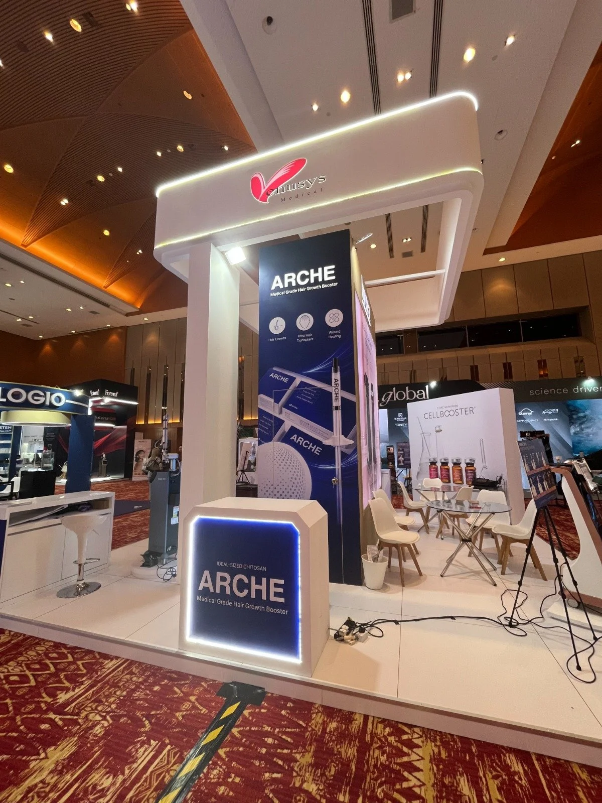 Booth at a trade show promoting ARCHE medical-grade hair growth booster. The booth has a vertical sign with product details, seating area with white chairs and glass table, and displays of medical bottles, set against a patterned carpet and large con