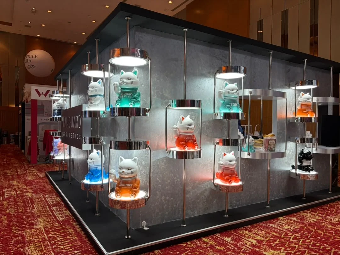 Display of illuminated cat figurines in glass cases at an exhibition or store, with red, green, blue, and black color accents, on a metallic stand with a patterned red carpet background.