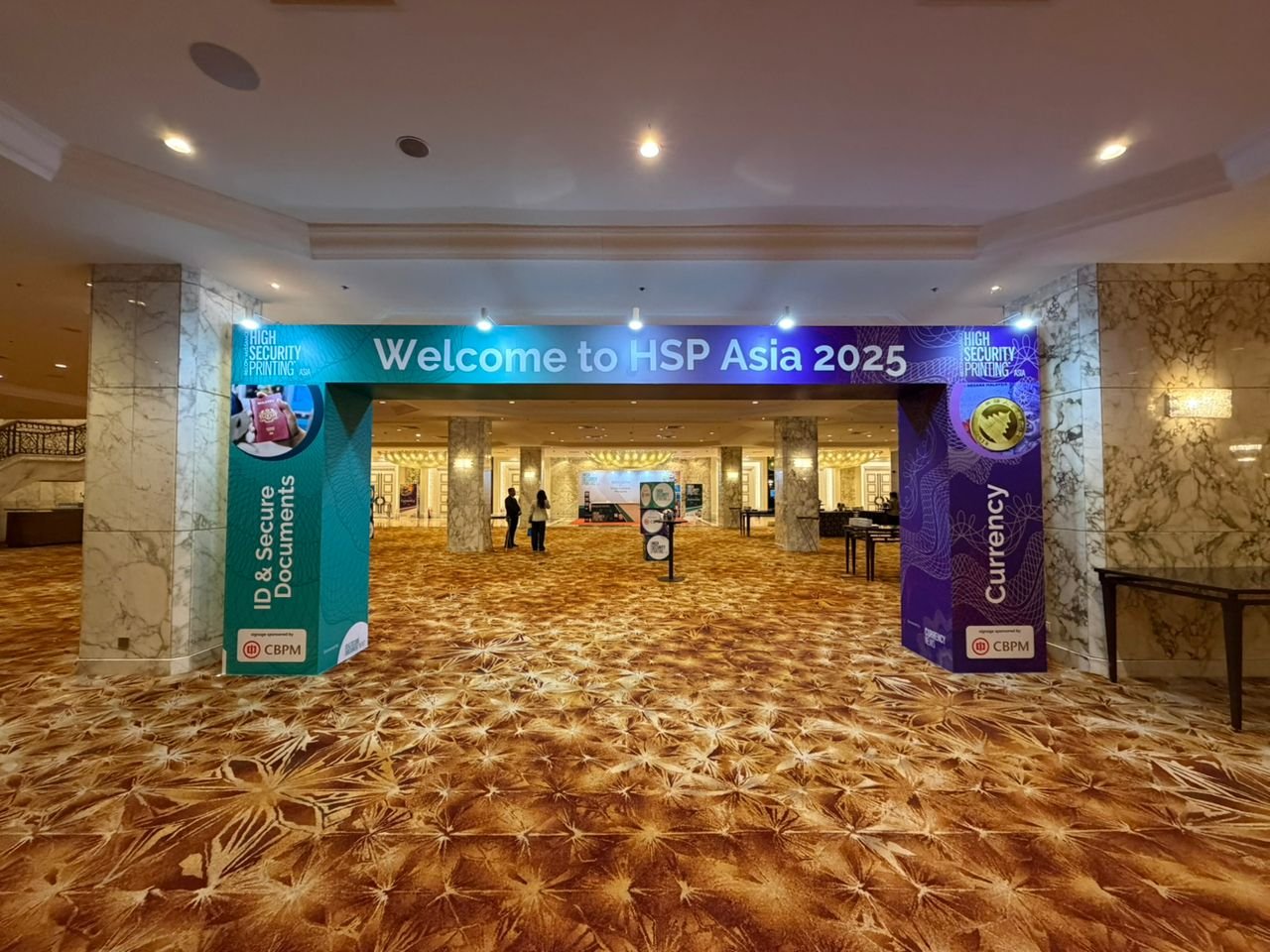 Entrance to the HSP Asia 2025 conference with a large blue and purple banner displaying the event name, welcoming attendees, and highlighting themes of high security printing, ID and secure documents, and currency.