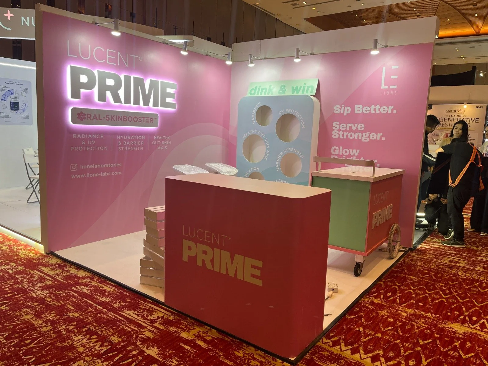 Exhibit booth for Lucent Prime with pink and green color scheme, promotional signage emphasizing skin protection and hydration, and a small mobi cart, set up at a trade show.