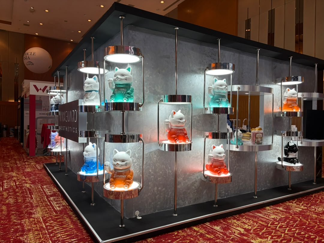 Display of colorful Maneki-neko cat figurines illuminated under lights on a modern metallic wall at an indoor event.