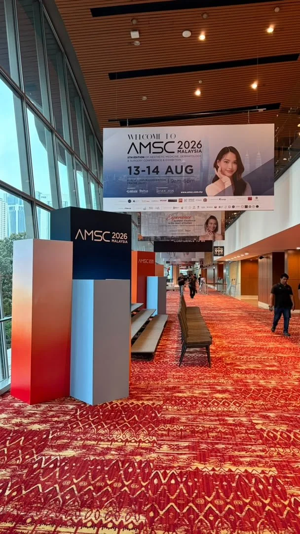 Conference hallway with red patterned carpet, large banners advertising AMSC 2026 Malaysia, and a few people walking. Side benches and a glass wall on the left.