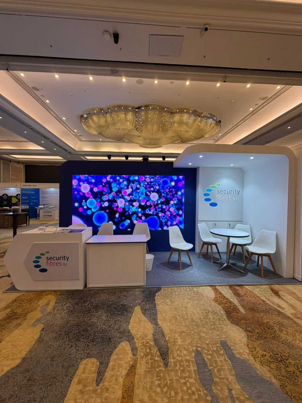 Exhibition booth for Security Fibres Ltd with a large digital screen displaying colorful bubbles, white chairs around small round tables, and a white counter with the company's logo.