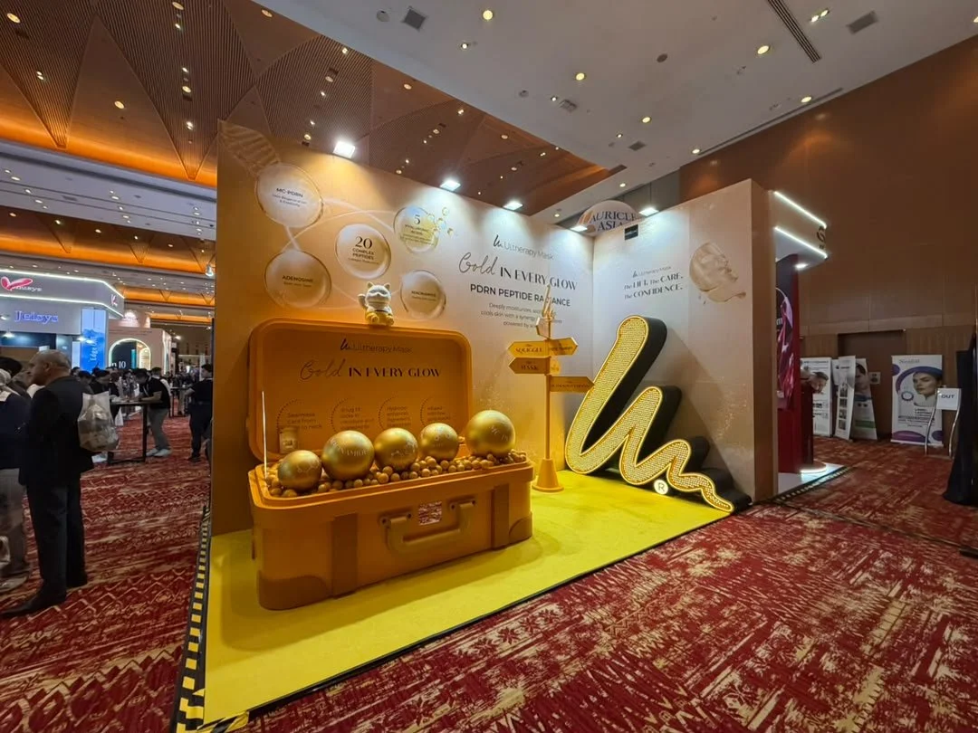 Exhibition booth with a yellow and white color theme promoting skincare products, featuring gold spheres, a large illuminated 'W' sign, and signs pointing to different product areas at a trade show.