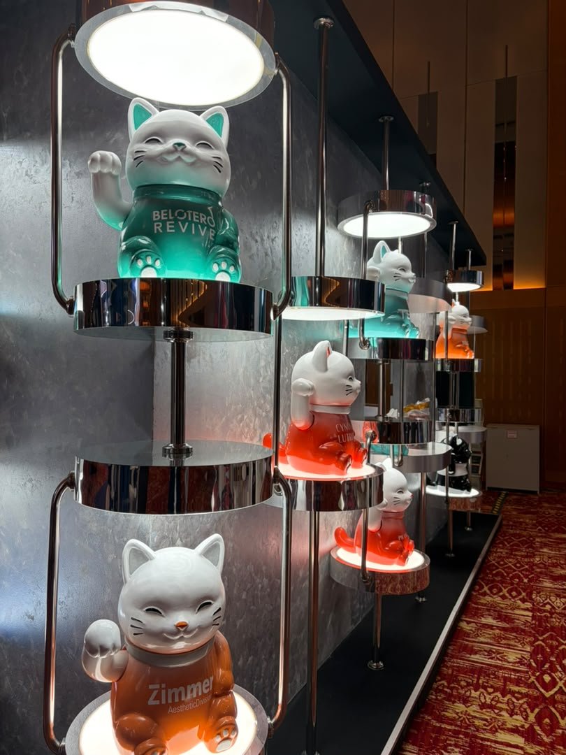 Display of illuminated maneki-neko figurines on a multi-tiered chrome stand. The figurines are white with some wearing color clothing. Each figurine has a raised paw and a friendly facial expression. The scene appears to be in a retail or exhibition 