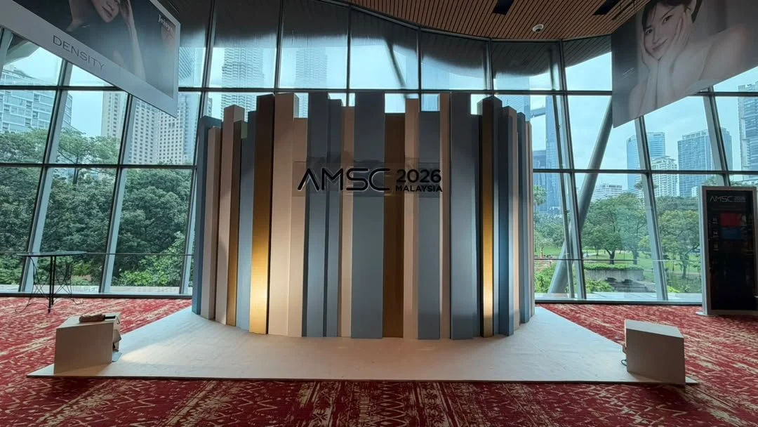 Modern conference or event stage with a circular design, featuring vertical panel decorations in gold, silver, and black. The stage has a sign reading "AMSC 2026 Malaysia". Large glass windows with cityscape and green outdoor scenery in the backgroun