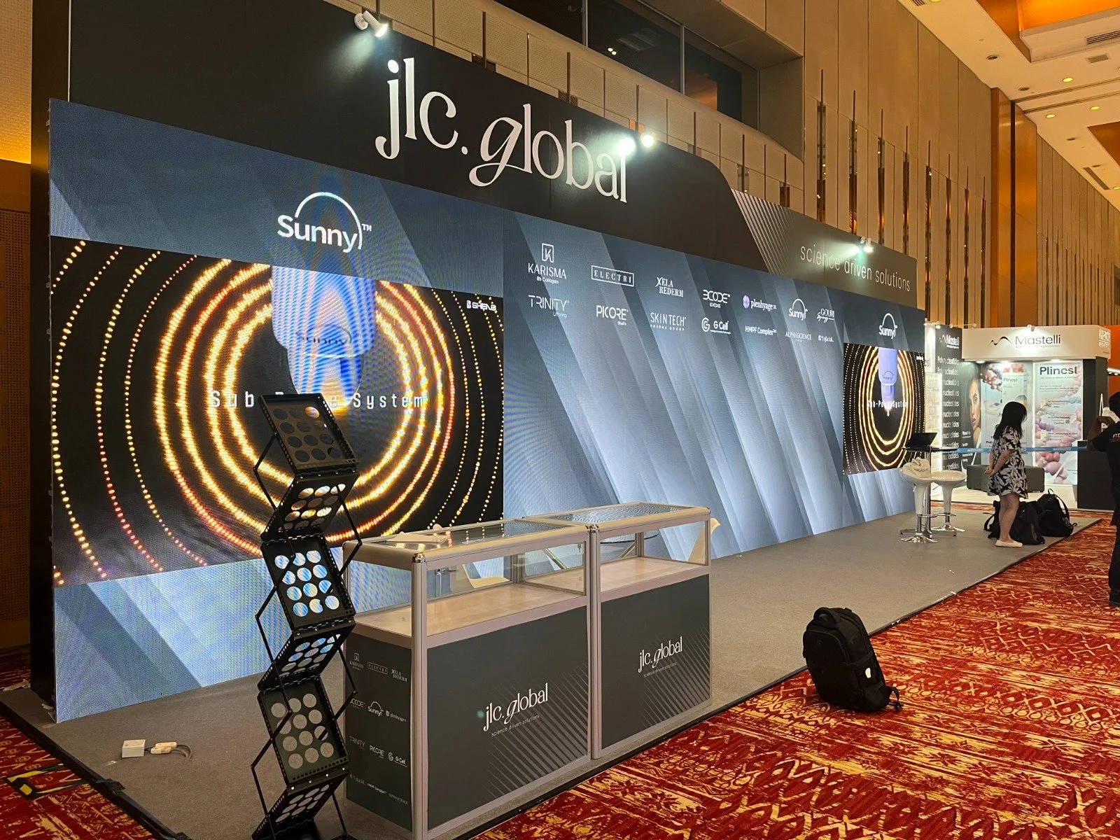 Trade show booth for jlc.global at a conference with digital screens, seating, and attendees, in a hotel conference hall with patterned red carpet.