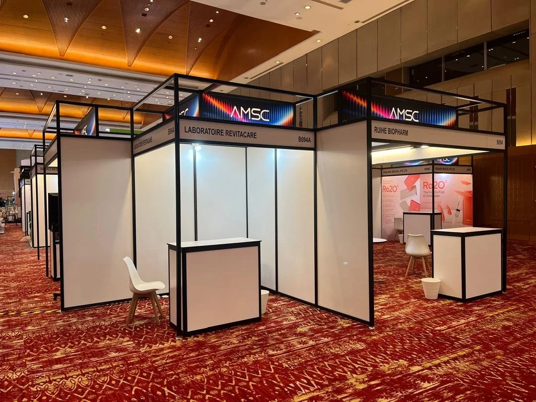 Conference exhibition booths with white walls and black frames at an event, with chairs and counters, and digital screens displaying colorful graphics and text.