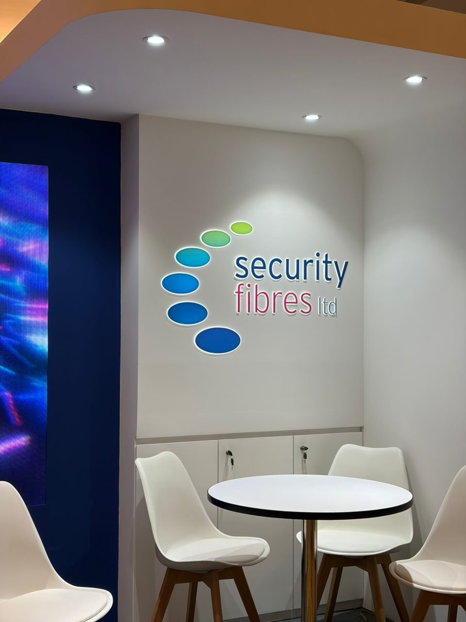 Interior of an office or conference room with a round white table, four white chairs with wooden legs, a white wall with a company logo, and ceiling lights.