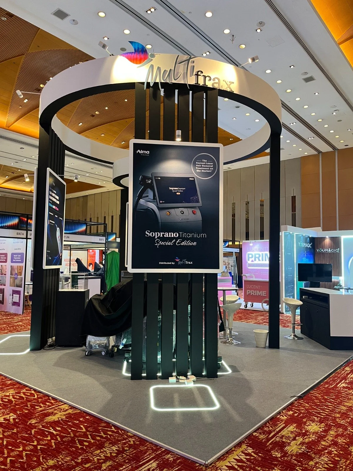 Exhibition booth at a convention center showcasing the Soprano Titanium Special Edition device by Muz Trax with informational posters and seating, under a high ceiling with ceiling lights.