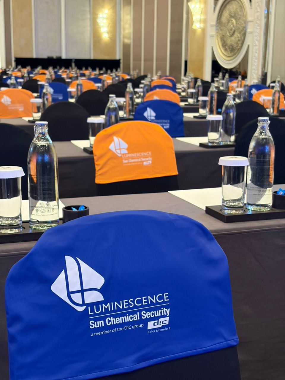 A conference room set up with tables and chairs covered in blue and orange chair covers, featuring the Luminescence Sun Chemical Security logo; water bottles and glasses placed on the tables.