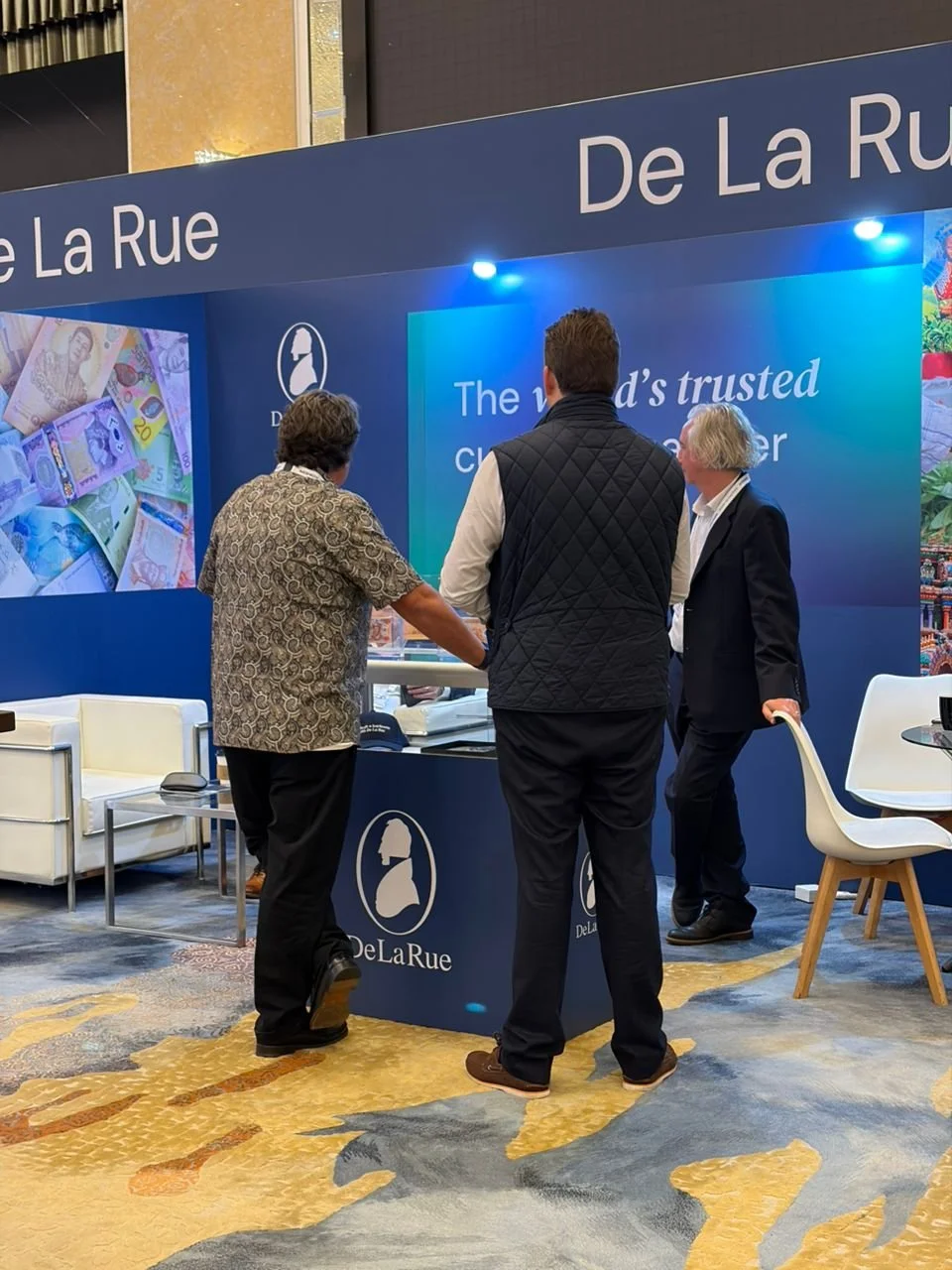 Three men are standing at a booth with the branding 'De La Rue' at a conference or exhibit. They are discussing; one man is facing a display screen, while two others are engaged in conversation. The booth has a blue backdrop, a large screen, and a pr