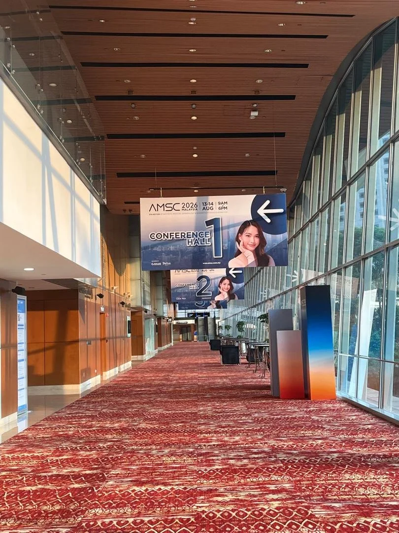 Long corridor with large glass windows on right, wooden ceiling, red patterned carpet, and conference hall signs for AMSA 2026 Malaysia, with white arrows pointing left, hanging from the ceiling.