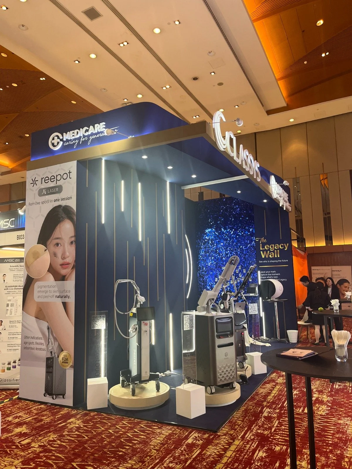A booth exhibit at a conference showcasing medical or cosmetic laser equipment, with a large poster of a woman with clear skin, and several high-tech devices on display.