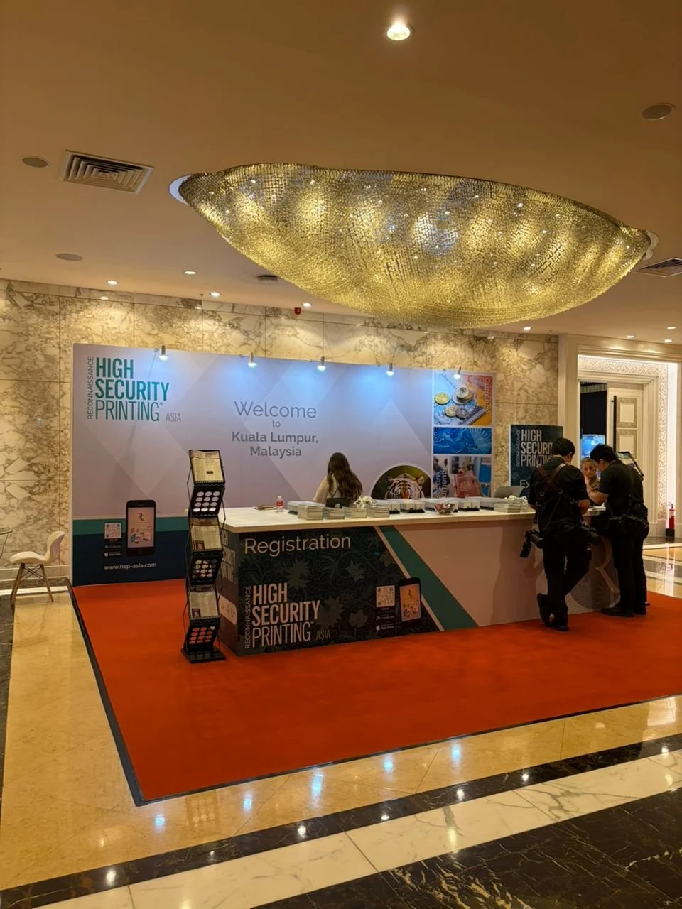 Registration desk at a high security printing conference in Kuala Lumpur, Malaysia, with a large chandelier overhead, and staff and attendees interacting.