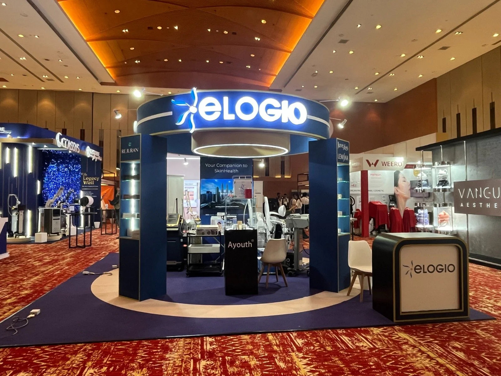 Exhibition booth for Elogio at a trade show, with illuminated Elogio sign, medical devices, and other brand displays, in a large conference hall with patterned red carpet.