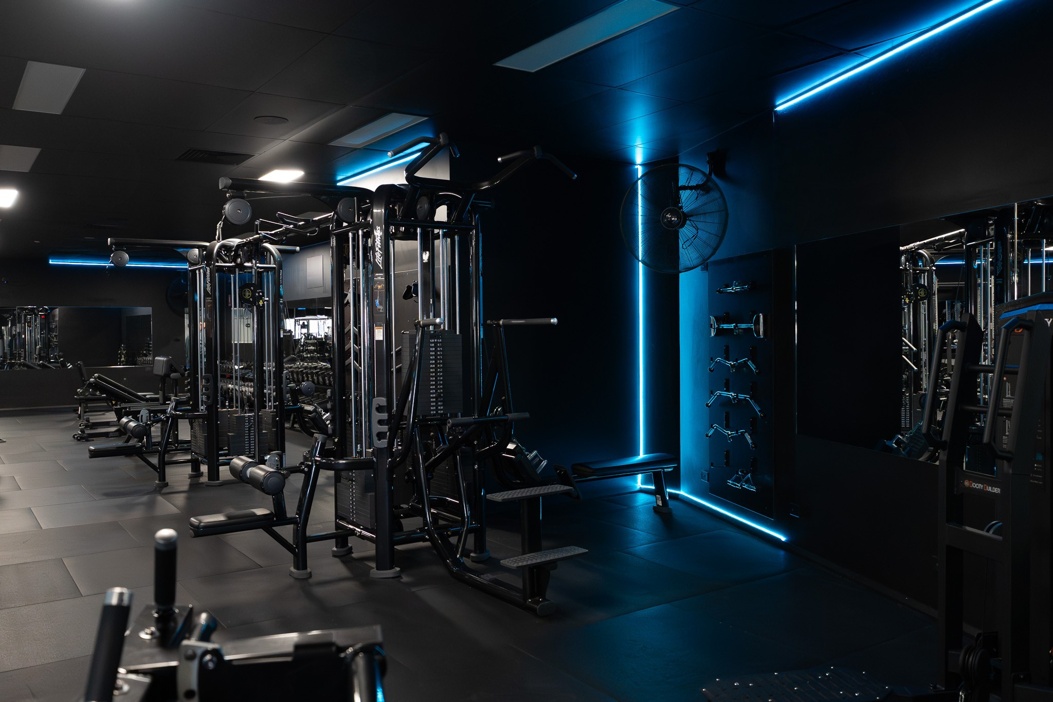 A dark gym room illuminated with blue LED lighting, featuring various exercise machines including weightlifting equipment and mirrors on the walls.