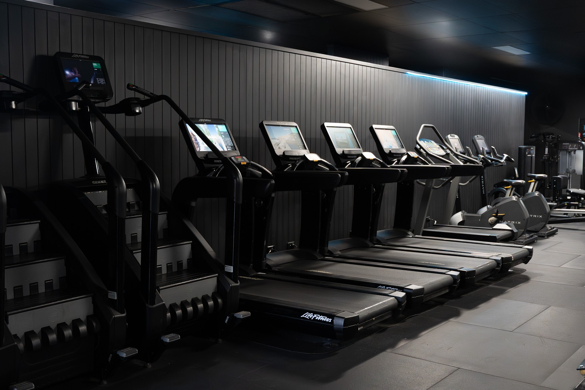 Row of exercise machines including treadmills in a dimly lit gym with dark walls and ceiling.