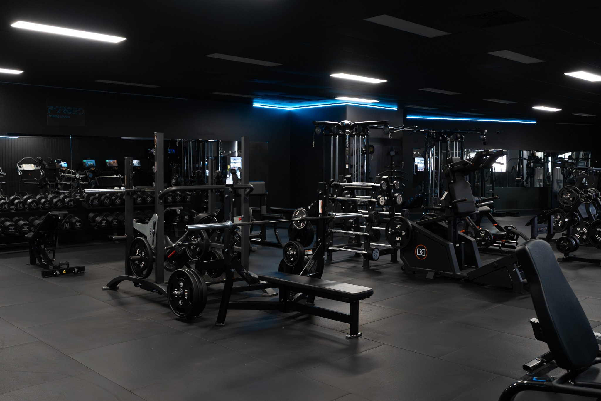Interior of a modern gym with various strength training equipment including dumbbells, weight benches, and machines, with dark walls and ceiling, illuminated by bright overhead lights and blue accent lighting.