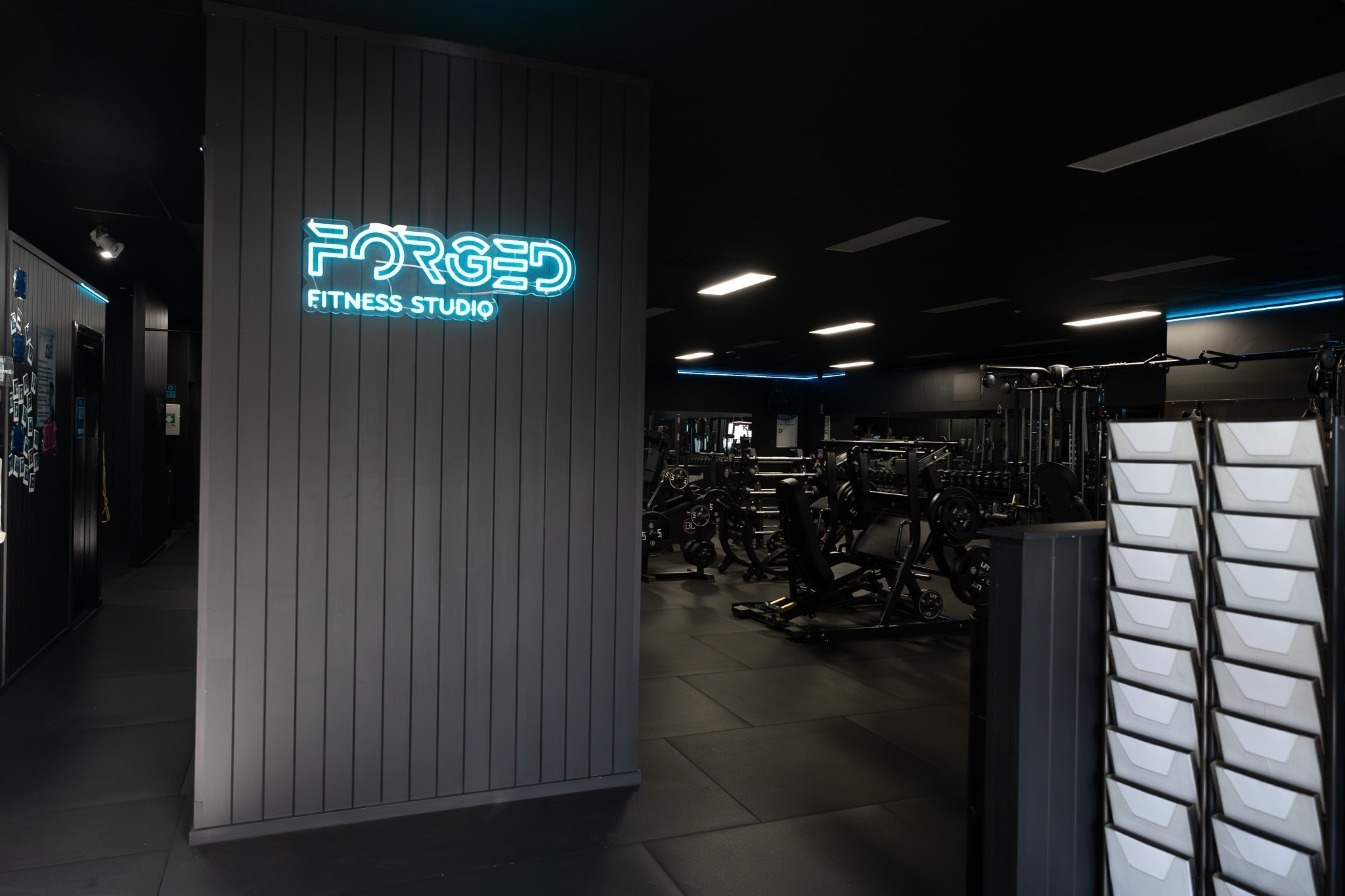 Interior of a fitness studio with gym equipment and a illuminated sign that reads 'FORGED FITNESS STUDIO' on a dark wall.