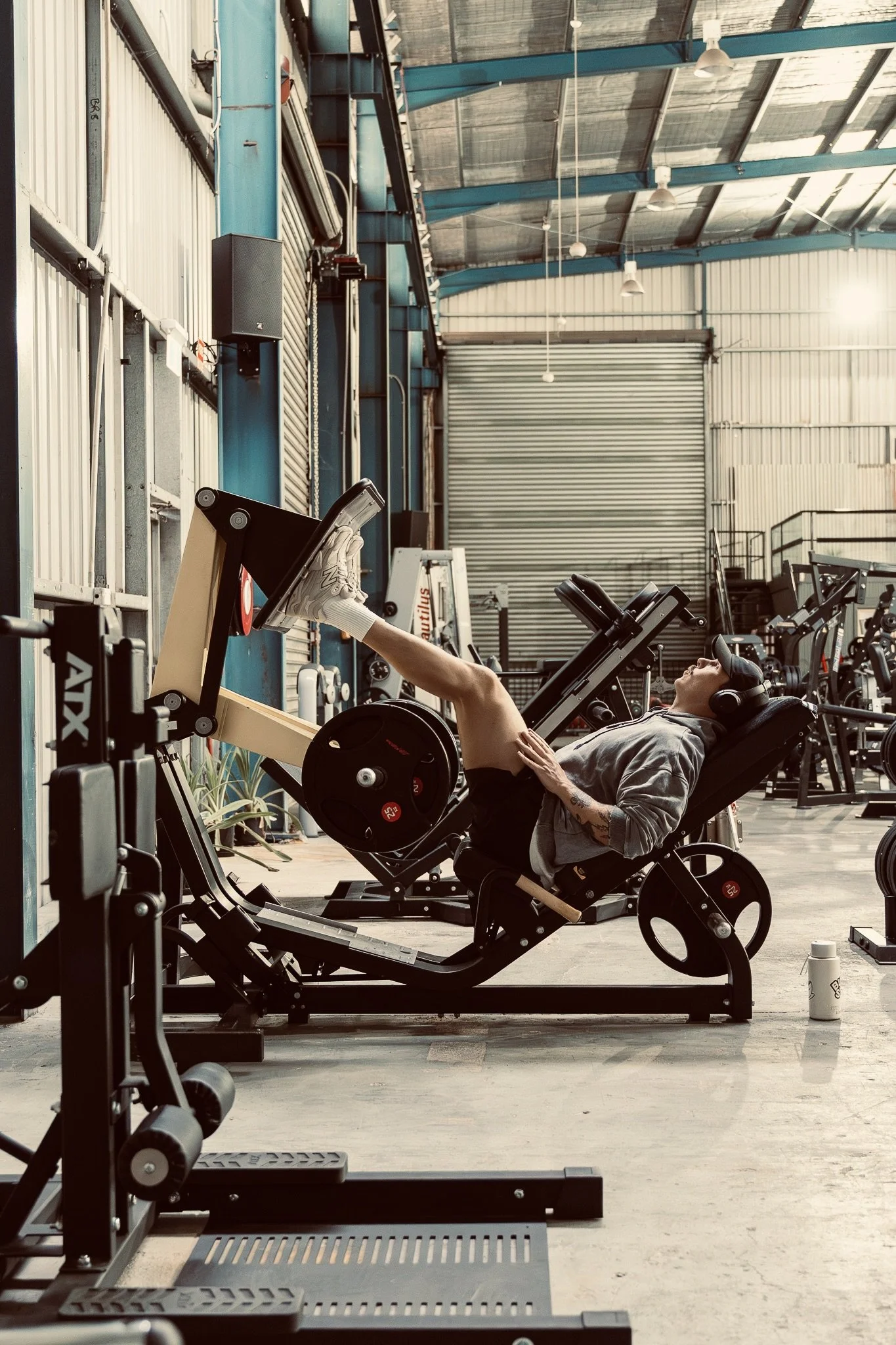 A person lying on a workout machine in a gym, lifting their legs with weights attached to their ankles. The gym has a high ceiling, metal walls, and various exercise equipment.