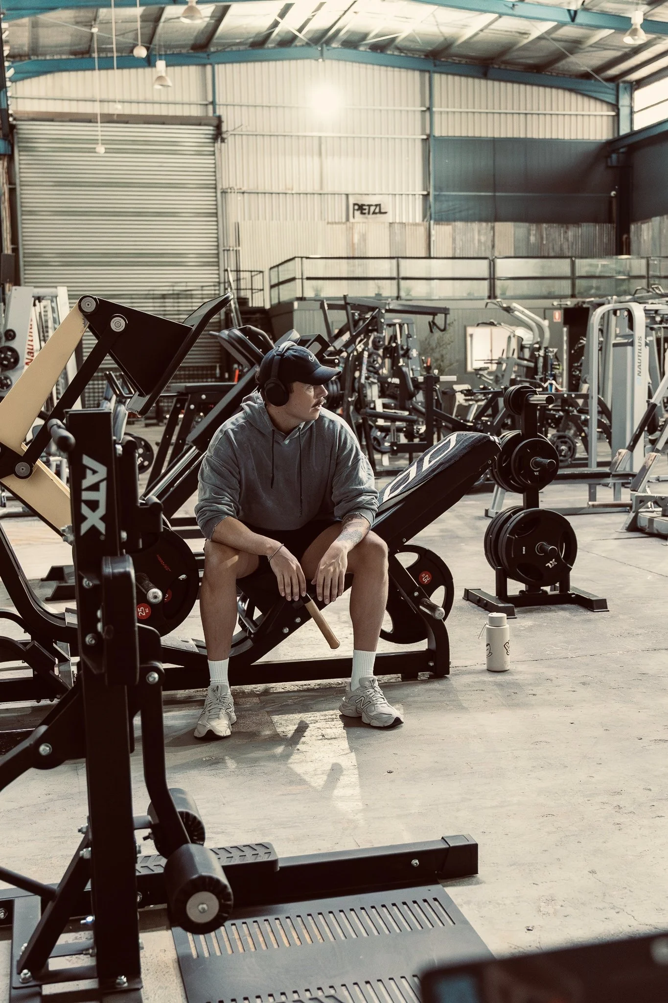 A young man wearing a gray hoodie, white shorts, and a baseball cap sits on a workout bench in a gym, wearing headphones and looking to his side. The gym is spacious with various exercise equipment and a metal wall with a sign that reads 'PETZL'. Sun