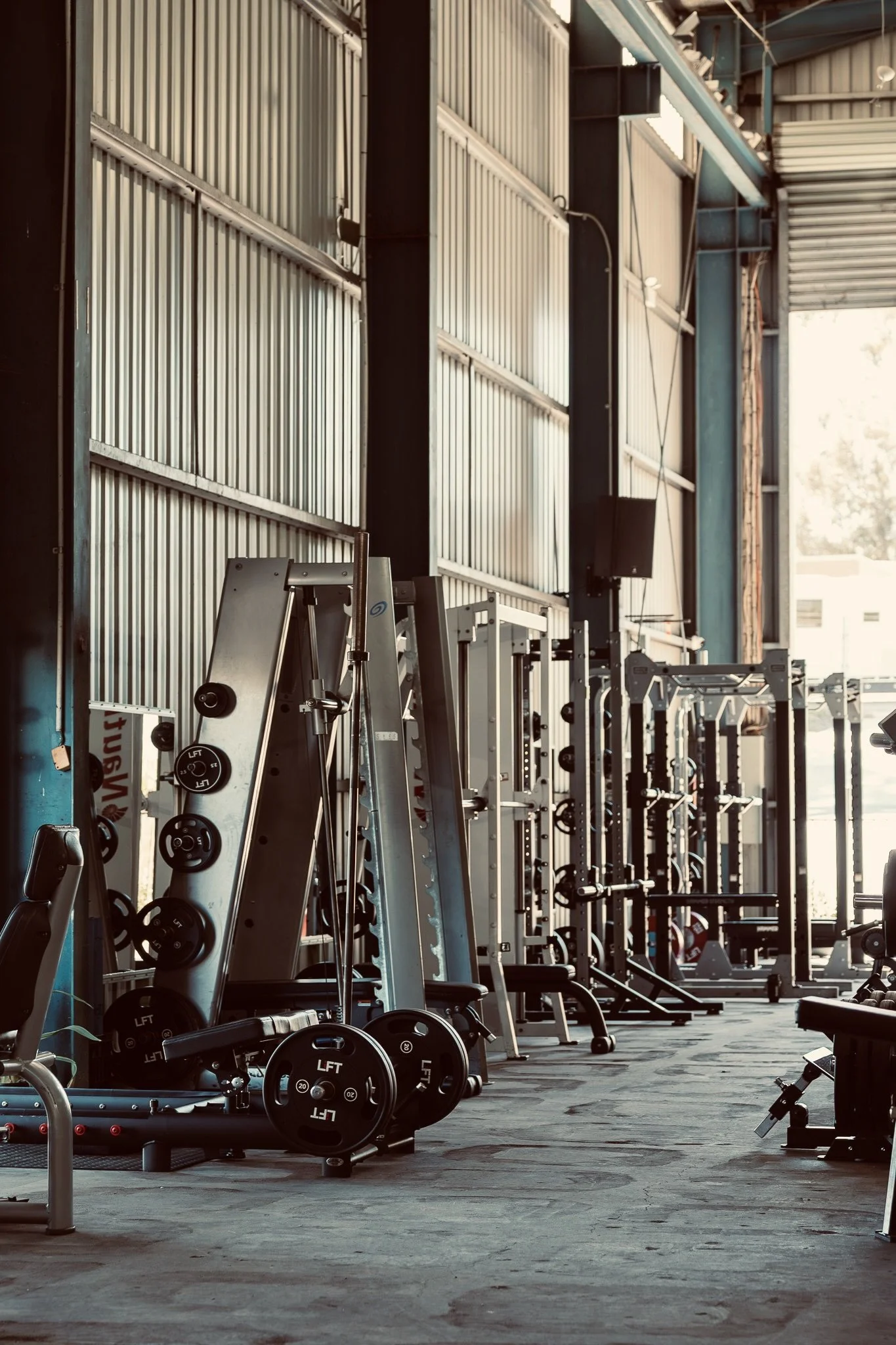 Empty gym with various weightlifting and workout equipment, including barbells, weight plates, and machines, in a well-lit industrial-style fitness center.