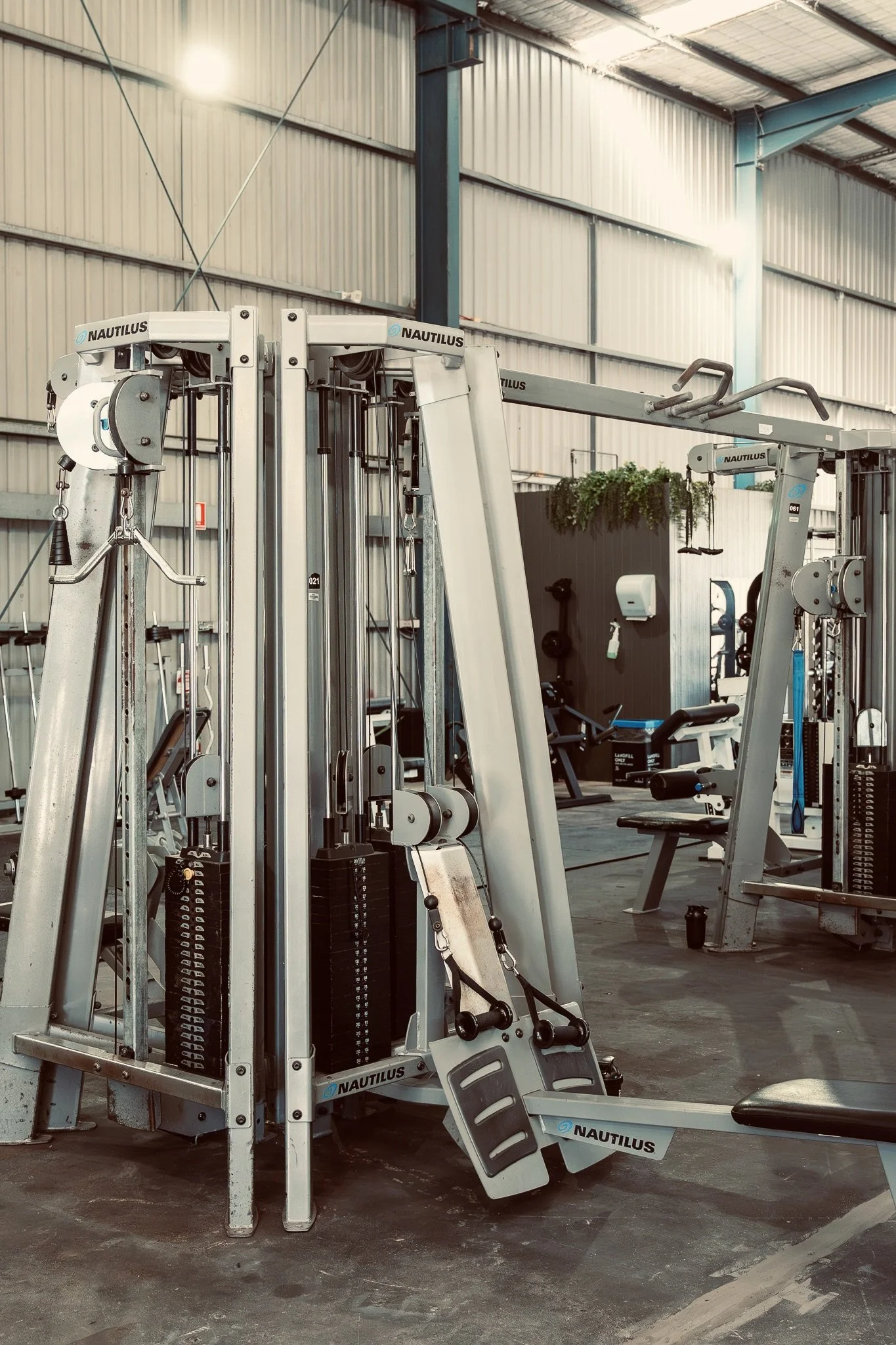 Inside a gym with various exercise machines and equipment, including a Nautilus resistance machine in the foreground, and a swing set attached to the machine. The gym has metal walls and a high ceiling with natural lighting.