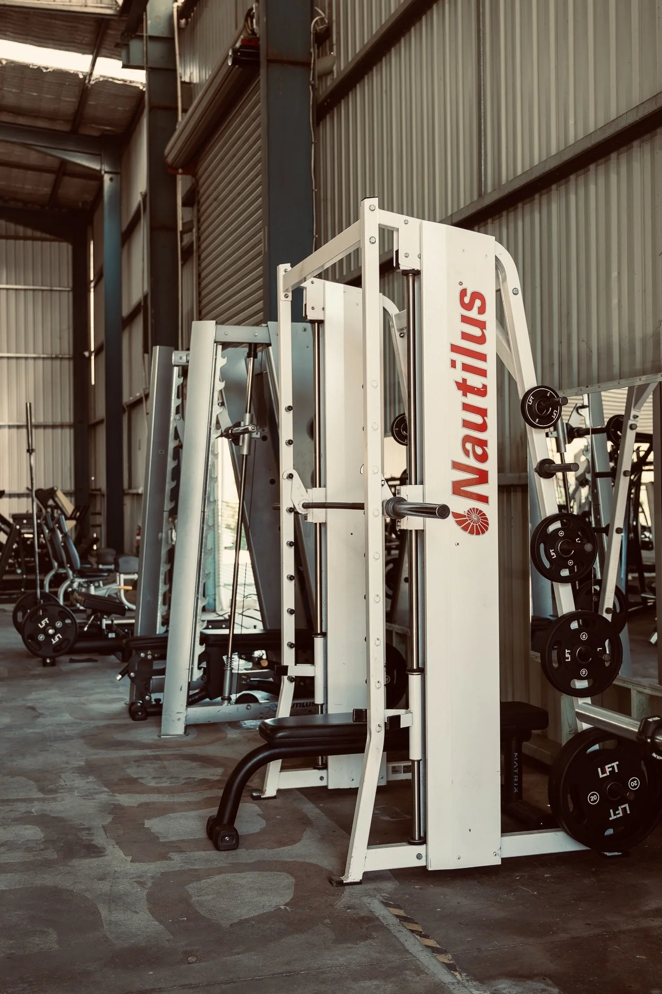 Empty gym with weightlifting equipment, including barbells and weight plates, in a spacious industrial-style workout area with metal walls and flooring.