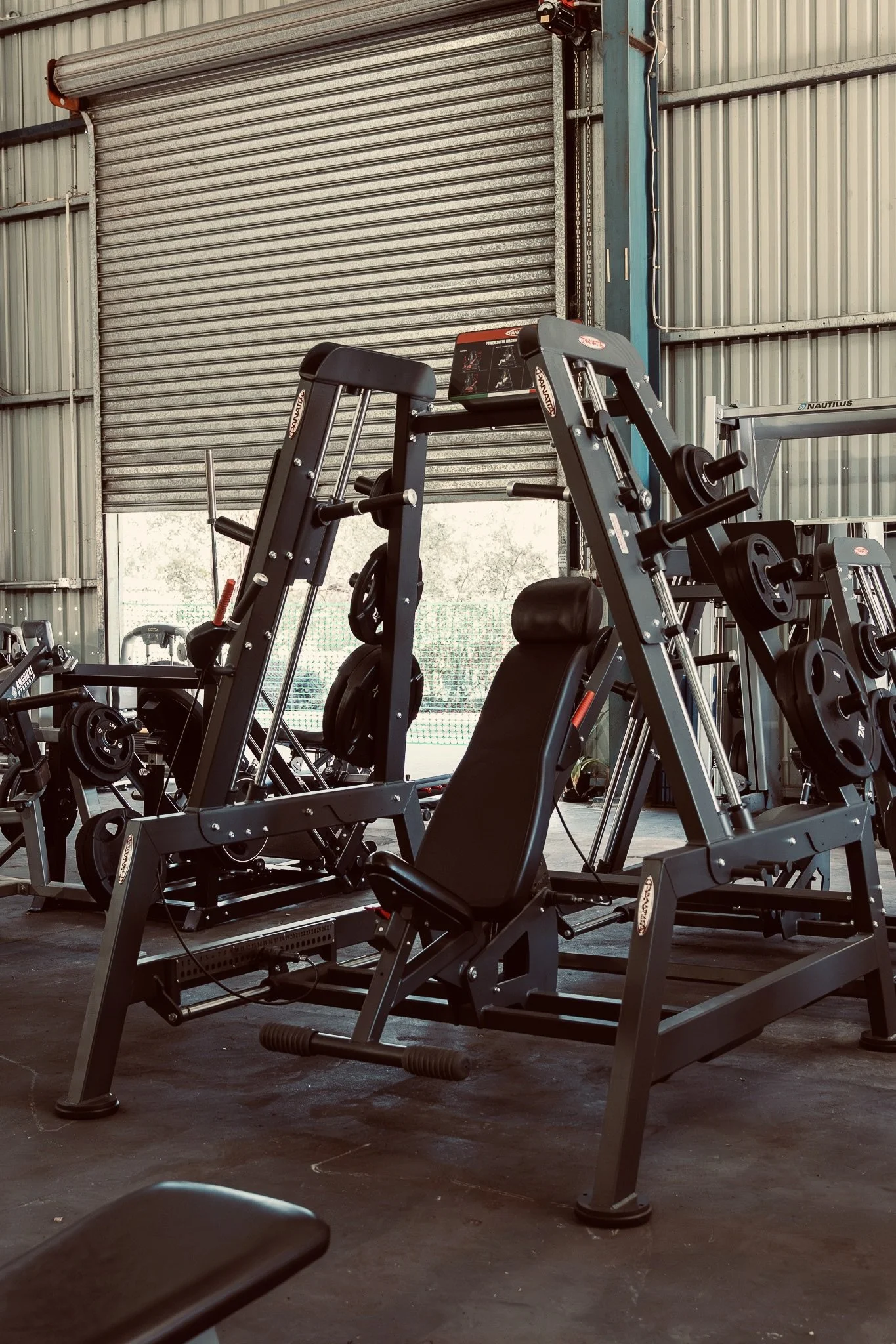 Gym with exercise equipment, including weight machines and a treadmill, inside a warehouse-like space with metal walls and a large roller door.