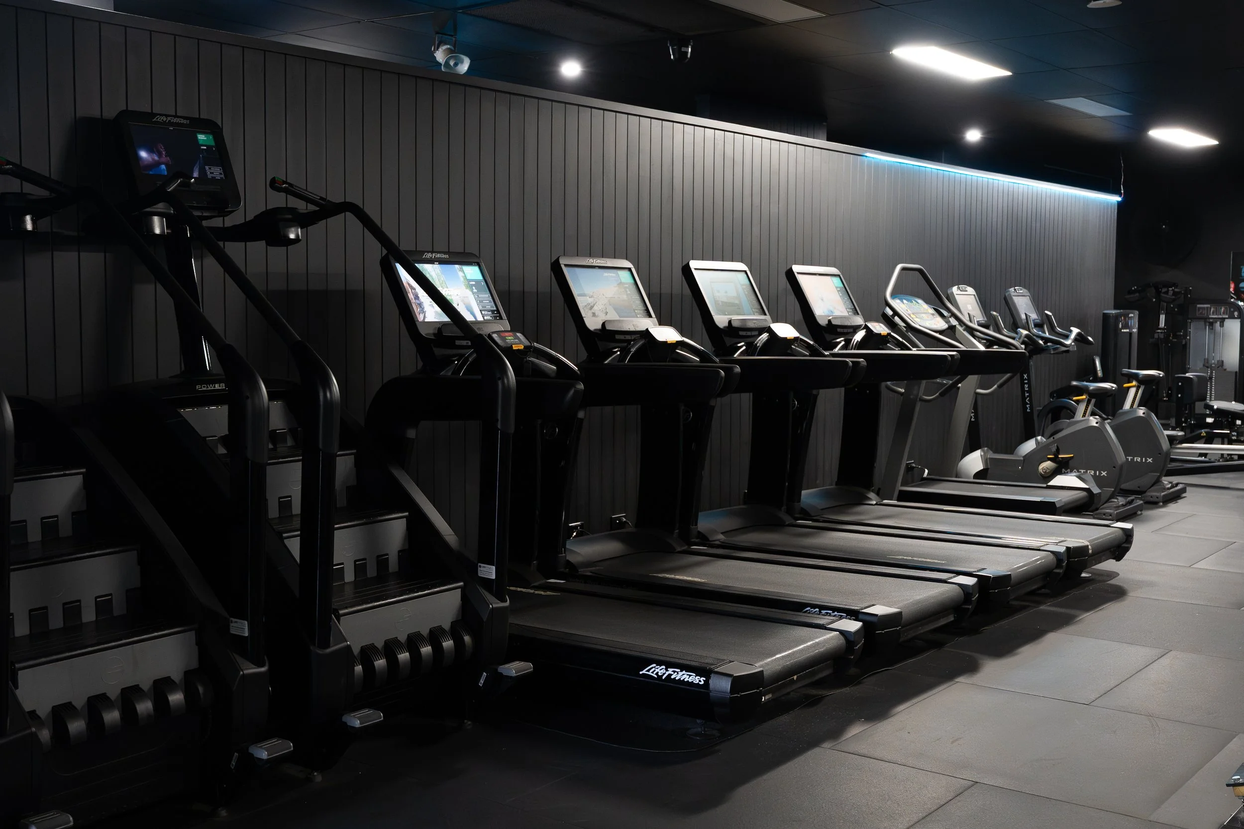 Row of treadmills and elliptical machines in a gym with dark walls and ceiling, illuminated by ceiling lights.