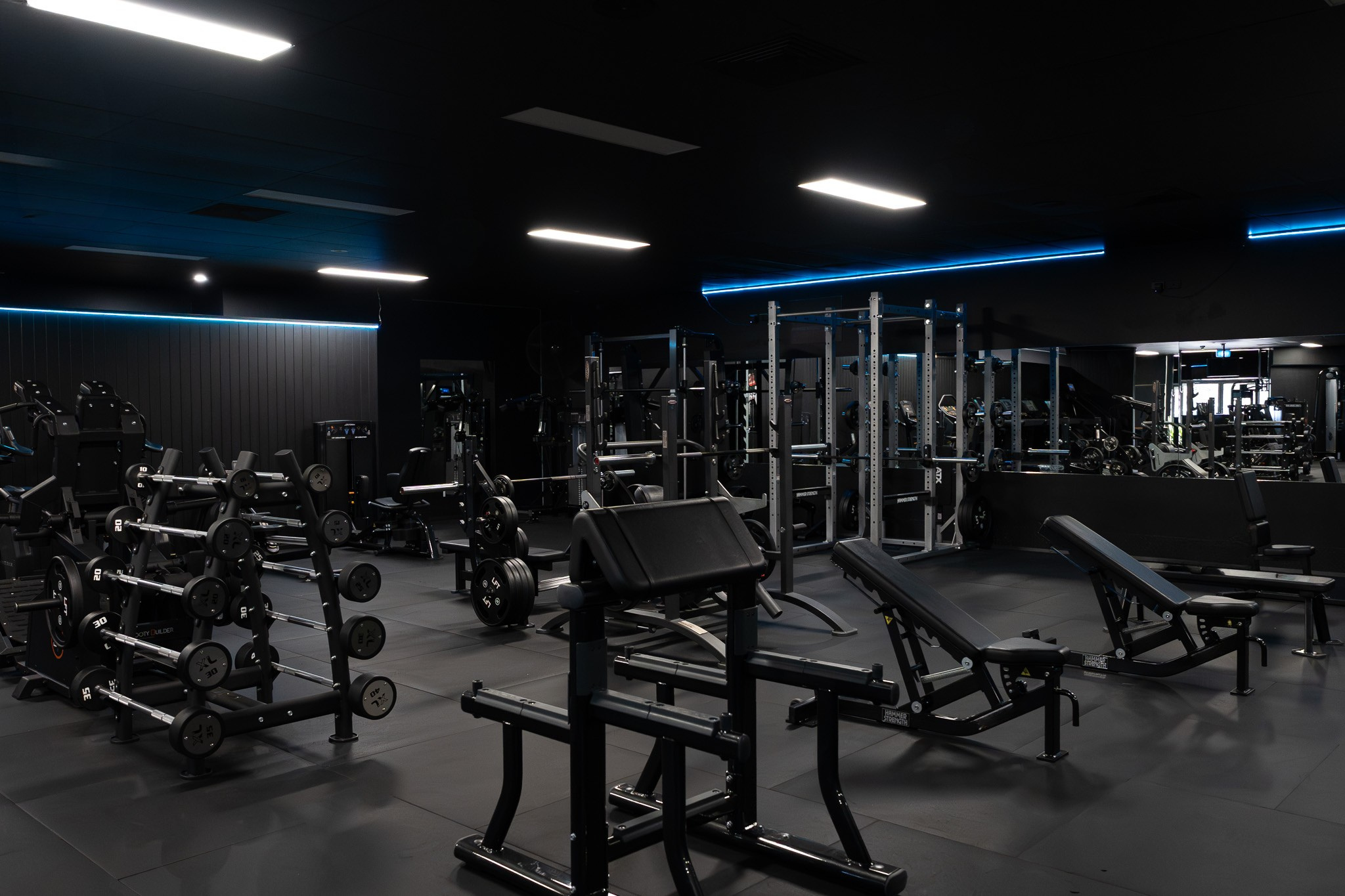 Interior view of a modern gym with various weightlifting equipment, including dumbbells, barbells, and weight machines, with black walls and ceiling, illuminated by bright overhead lights and blue LED lighting.