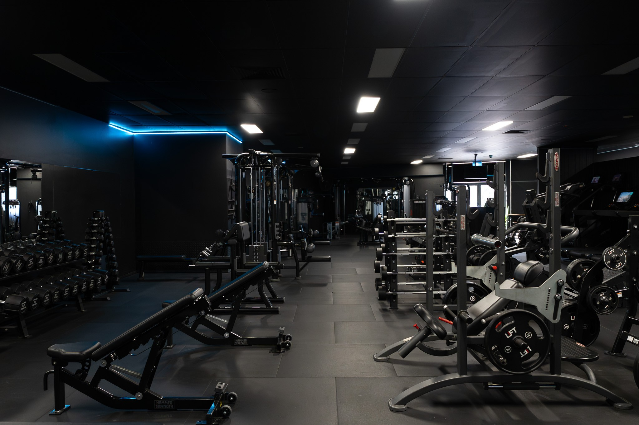 Interior of a modern, dimly lit gym with various types of workout equipment including weights, benches, and machines, illuminated by ceiling lights and blue accent lighting.