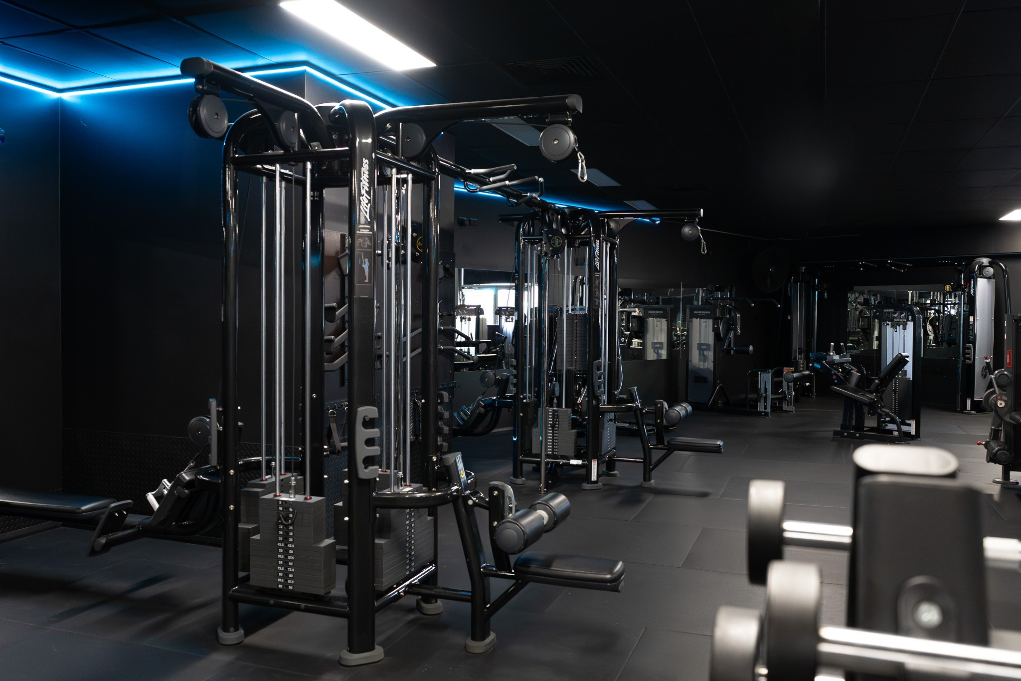 Interior view of a modern gym with various strength training equipment and black walls with blue accent lighting.